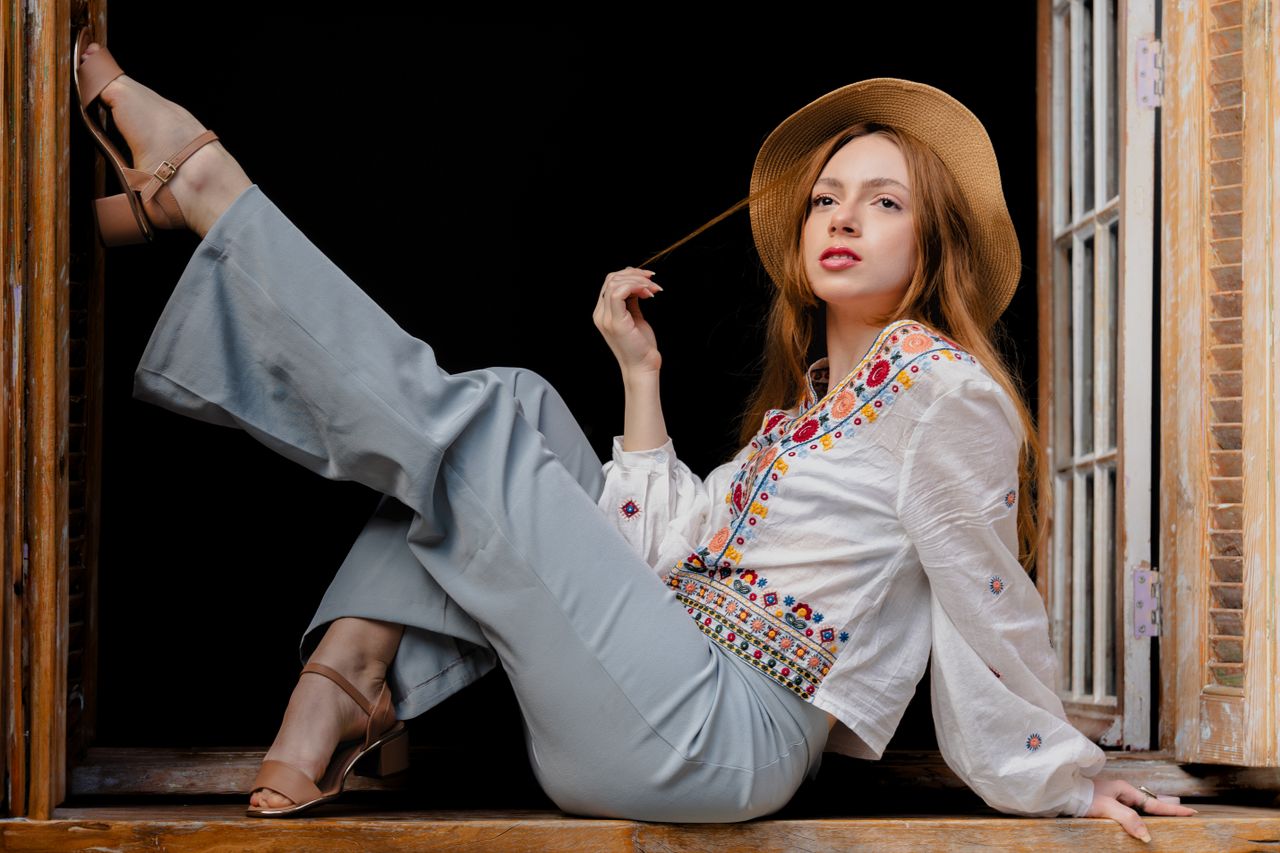 creative-03 Young woman posing elegantly by a windowsill, wearing fashionable embroidered blouse and hat.